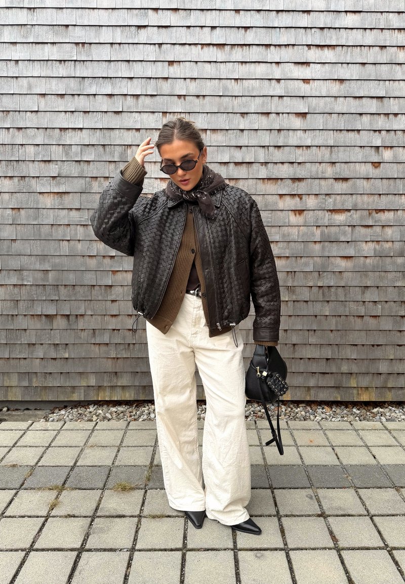 Personne portant des lunettes de soleil, une veste en cuir tressé foncé, une chemise marron, un pantalon large blanc, des chaussures noires pointues, tenant un sac noir clouté près d'un mur en bois.