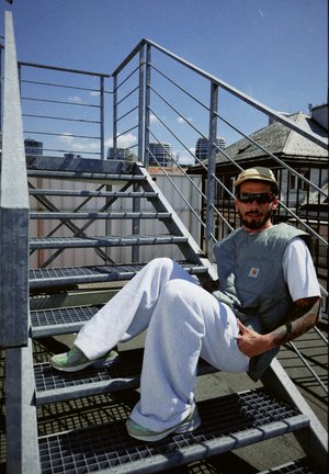 Escaleras de metal grises con una persona sentada, vestida con un chaleco gris, pantalones blancos y zapatos con detalles verdes. Cielo azul de fondo.