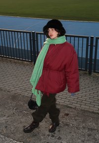 Κόκκινο μπουφάν με ζώνη στη μέση, μοντέρνο πράσινο υφασμάτινο κασκόλ, σκούρο πράσινο παντελόνι και μαύρες ankle μπότες. Μαύρο καπέλο από γούνα και μικρή μαύρη τσάντα.