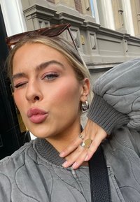 Graue gesteppte Bomberjacke, silberne Ohrringe, Ringe an den Fingern, braune Sonnenbrille auf dem Kopf, leichtes Zwinkern, dezentes Make-up mit rosa Tönen, neutraler Hintergrund.