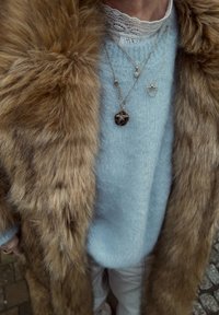 Fuskepelsfrakke i brun, lyseblå strikket sweater, lagdelte guldkæder med vedhæng. Blød tekstur og neutral farvepalette.