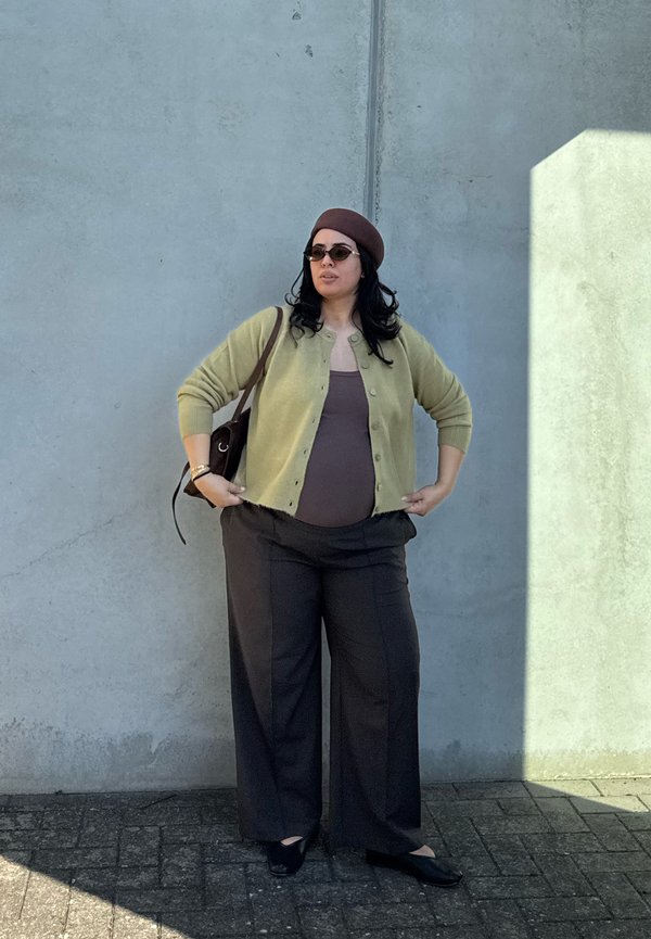 Mujer embarazada con gafas de sol, boina, cárdigan verde y pantalones anchos oscuros, de pie contra una pared gris con sombra a la derecha.