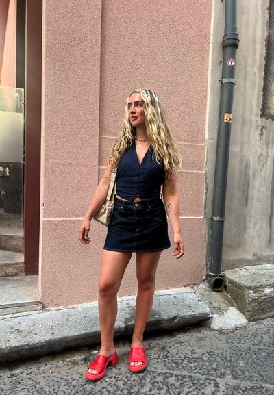 Top corto azul oscuro combinado con una minifalda de mezclilla oscura. La persona lleva sandalias rojas y sostiene un bolso beige. Fondo urbano texturizado.