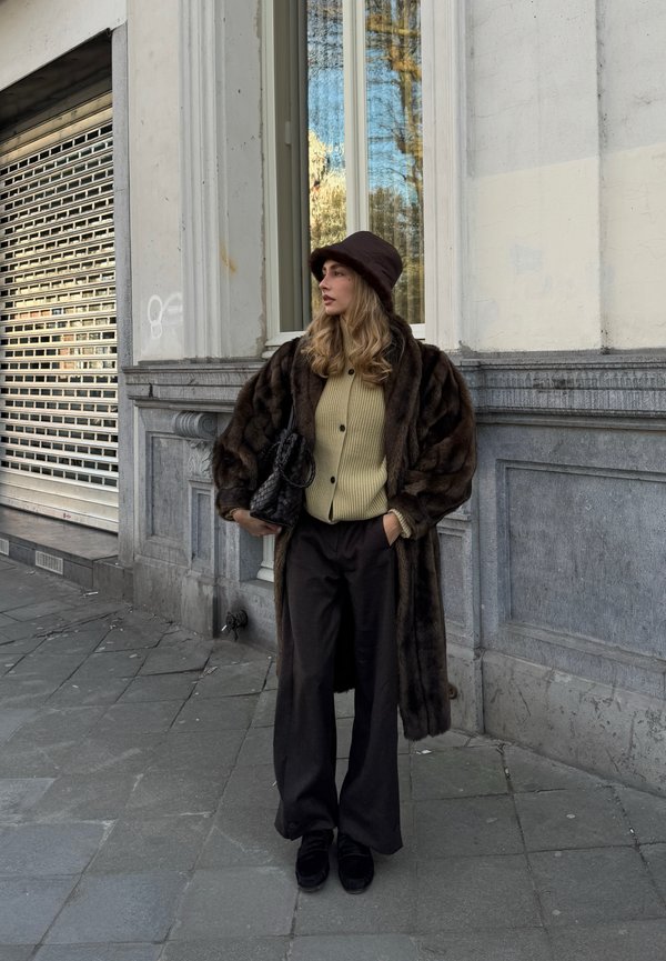 Femeie purtând o haină de blană închisă la culoare, pălărie tip găleată, pulover bej cu nasturi, pantaloni largi negri și ținând o geantă neagră țesută, stând lângă o clădire.