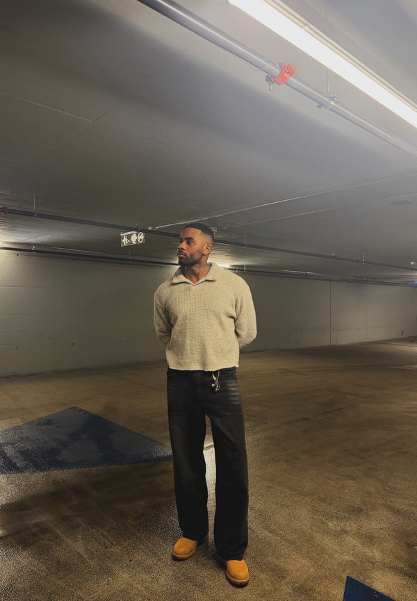 Man in beige jumper, black trousers, and tan boots standing with hands behind back in a dimly lit indoor car park.