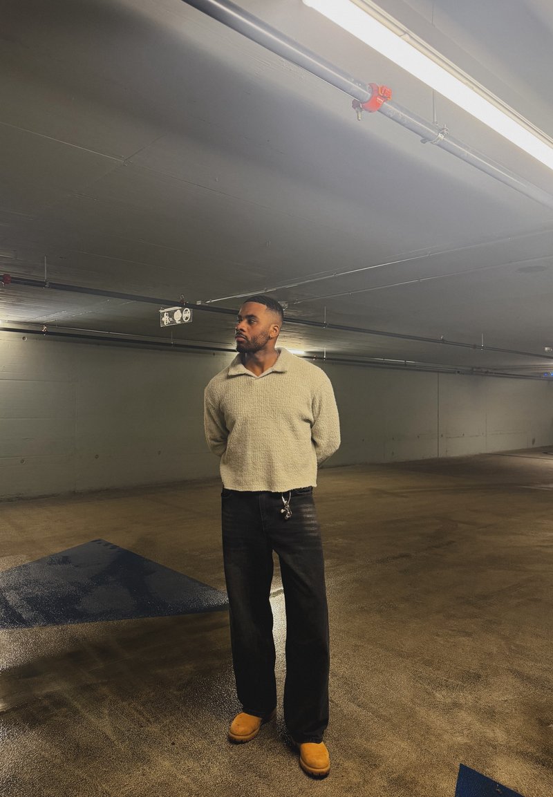 Homme en pull beige, pantalon noir et bottes couleur sable, debout les mains derrière le dos dans un parking intérieur faiblement éclairé.