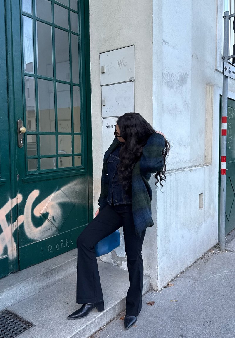 Γυναίκα με μακριά σκουρόχρωμα μαλλιά, ντυμένη με μαύρο παντελόνι και σακάκι, που στρώνει τα μαλλιά της κοντά σε μια πράσινη πόρτα με γκράφιτι σε πεζοδρόμιο της πόλης.