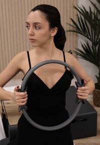 Anneau de Pilates gris, avec poignées texturées et design circulaire. L'utilisateur tient l'anneau dans un cadre sportif avec des plantes d'intérieur et du mobilier.