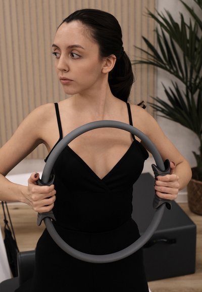 Anillo de Pilates en gris, con agarres texturizados y diseño circular. El usuario sostiene el anillo en un entorno de fitness con plantas de interior y mobiliario.