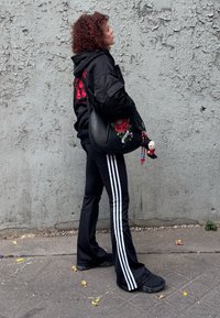 Melna bumbvedēja jaka ar sarkanu grafisku dizainu, melnas izplatītas bikses ar baltām svītrām, melni apavi un soma ar ziedu izšuvumu.