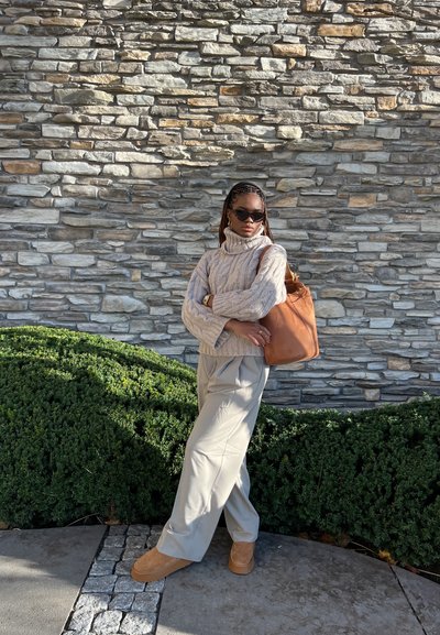 Jersey de cuello alto beige, pantalones anchos crema, bolso marrón, calzado marrón. De pie contra una pared de piedra con vegetación verde.