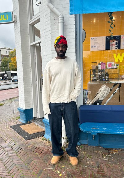 Suéter de punto blanco, oversized, con dobladillo deshilachado, jeans azul oscuro y botas marrones. Gorro colorido con patrón de hojas. De pie frente a un café.