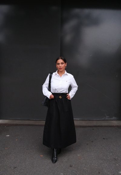 Camisa blanca de botones, falda negra de talle alto, botines negros con cordones, bolso negro, hebilla de cinturón dorada prominente, sobre un fondo gris.
