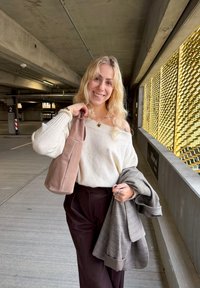 Een vrouw in een lichtbeige off-shoulder trui houdt een bruine tas en een grijs geruit blazer vast, poseert in een parkeergarage met patroonwanden.