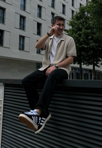 Camisa de punto beige de manga corta sobre una camiseta blanca, jeans negros y zapatillas negras con rayas blancas, sentado en un borde oscuro bajo.