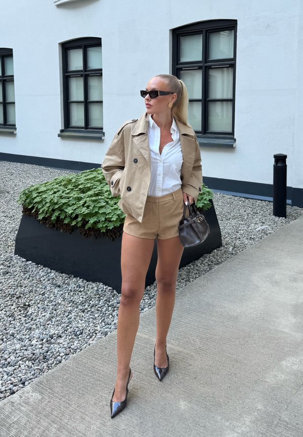 Frau in beigen Shorts und Jacke, weissem Hemd, schwarzen Absätzen, Sonnenbrille und Pferdeschwanz steht auf einem Betonweg neben einem modernen Gebäude und Grünfläche.