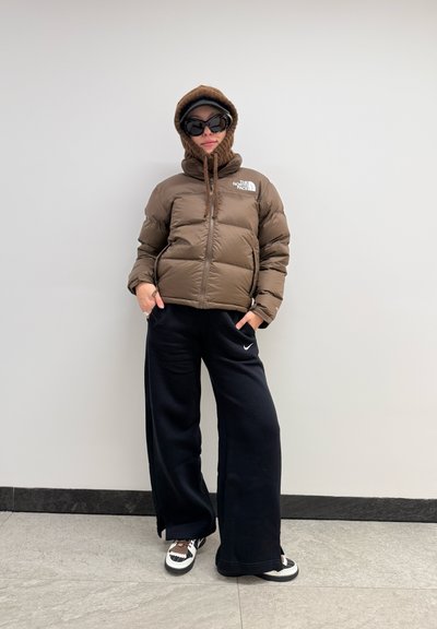 Chaqueta de abrigo marrón con capucha, pantalones de pierna ancha negros y zapatos en blanco y negro. Un par de gafas de sol y una capucha de punto realzan el conjunto.