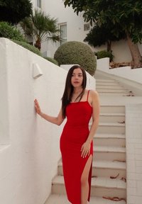 Rouge fitted dress with slender straps, a side slit, and a smooth texture. The model leans against a white wall next to stairs, surrounded by lush greenery.
