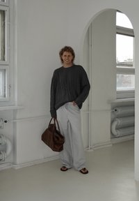 Suéter cinza de malha grossa, calças largas listradas, bolsa de couro castanho e chinelos. Ambiente interior com paredes brancas e grandes janelas.