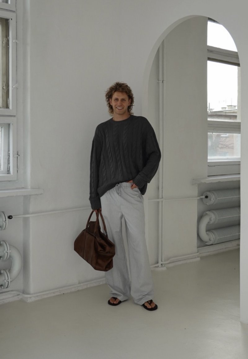 Grauer Kabelstrickpullover, gestreifte weite Hose, braune Ledertasche und Flip-Flops. Innenraum mit weißen Wänden und großen Fenstern.