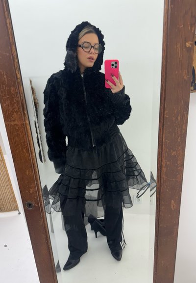 Chaqueta de imitación de piel negra con cremallera y capucha, falda negra de capas con volantes transparente, pantalones negros con rayas blancas y tacones puntiagudos negros.