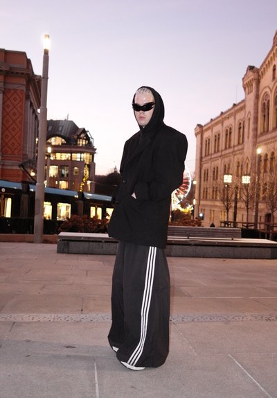 Absoluto abrigo negro de gran tamaño con capucha, pantalones negros con rayas blancas y gafas de sol. Entorno urbano, luz del atardecer, arquitectura moderna de fondo.