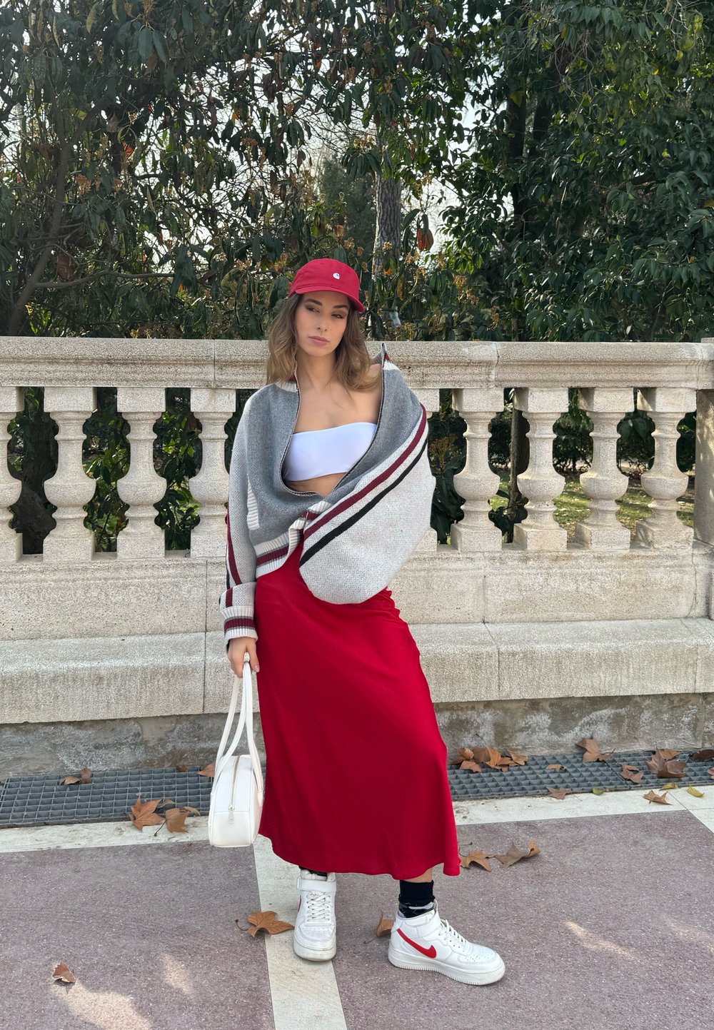 Grijze oversized trui met kastanjebruine strepen, witte crop top, rode maxi rok, witte handtas en witte sneakers met rode accenten. Natuurlijke omgeving.