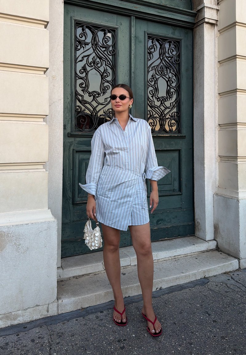 Femme en robe chemise rayée et sandales rouges se tient devant des portes doubles ornées vertes, tenant un petit sac à main blanc en forme de coquillage.