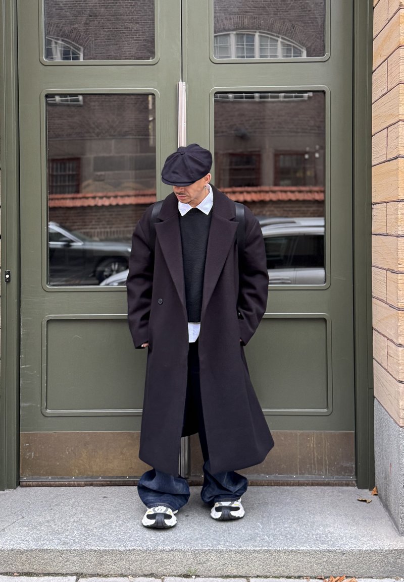 Lungo soprabito nero, lana strutturata, sovrapposto a un maglione scuro e a una camicia bianca. Jeans scuri, oversize, abbinati a sneakers bianche e nere.