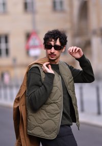 Olivovo zelená prešívaná vesta na tmavozelenom svetri, spárovaná so šedými nohavicami. Čierne slnečné okuliare, drží hnedú bundu cez jedno rameno.