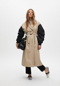 Trench coat beige con maniche nere, chiusura doppiopetto, cintura annodata e tasche laterali. Abbinato a una borsa nera e scarpe argento.