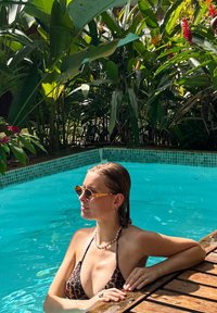 Bikini à imprimé léopard, collier de perles et lunettes de soleil avec accents orange. Femme près d'une piscine turquoise entourée de plantes vertes luxuriantes. Terrasse en bois visible.