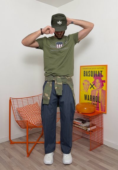 Hombre con camiseta verde, chaqueta de camuflaje atada a la cintura, pantalones de rayas finas y gorra, de pie cerca de una silla naranja y un cartel de Basquiat-Warhol.