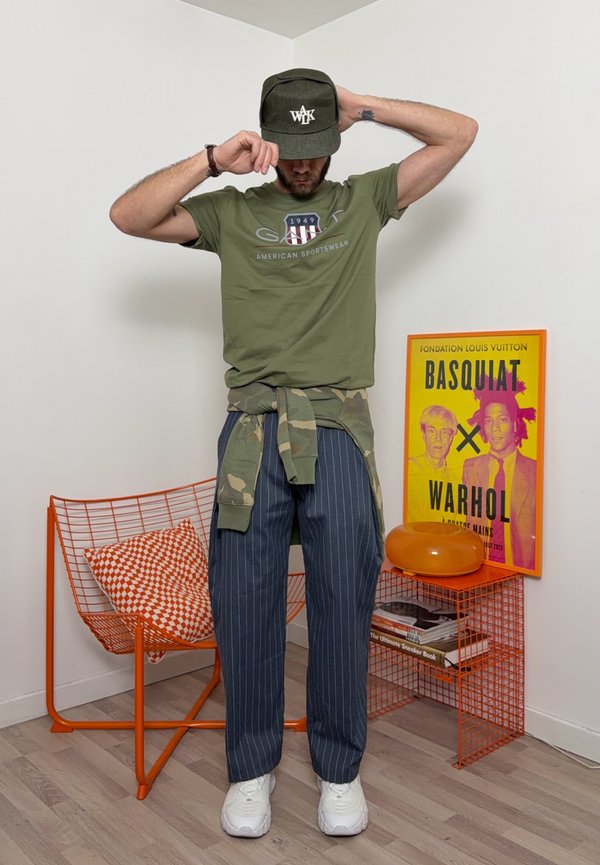 Homme portant un t-shirt vert, une veste camouflage attachée à la taille, un pantalon à fines rayures et une casquette, se tenant près d'une chaise orange et d'une affiche Basquiat-Warhol.