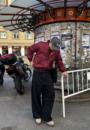 Persona con chaqueta granate y gorra azul marino ajustándose pantalones anchos negros, de pie junto a una motocicleta y un quiosco cubierto de graffiti en la acera de la ciudad.