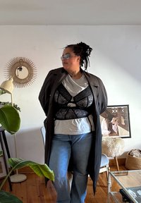 Une superposition de dentelle noire sur un corsage ajusté porté sur un t-shirt gris, associée à un jean flare en denim et un long manteau sombre. Une lumière douce illumine la scène.