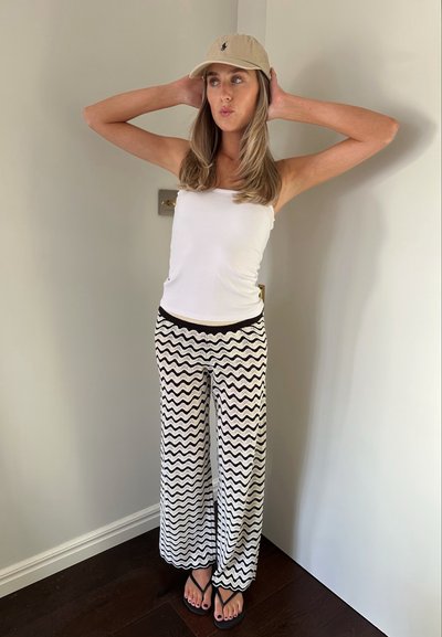 Top sin mangas blanco, pantalones de pierna ancha con patrón de zigzag en blanco y negro, gorra beige y chanclas negras sobre un suelo de madera contra una pared clara.