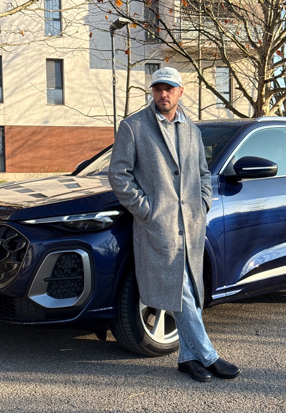 Un homme portant un manteau gris et une casquette se tient les mains dans les poches, appuyé contre l'avant d'un SUV bleu foncé sur une rue pavée.