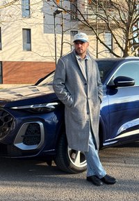 Un homme portant un manteau gris et une casquette se tient les mains dans les poches, appuyé contre l'avant d'un SUV bleu foncé sur une rue pavée.