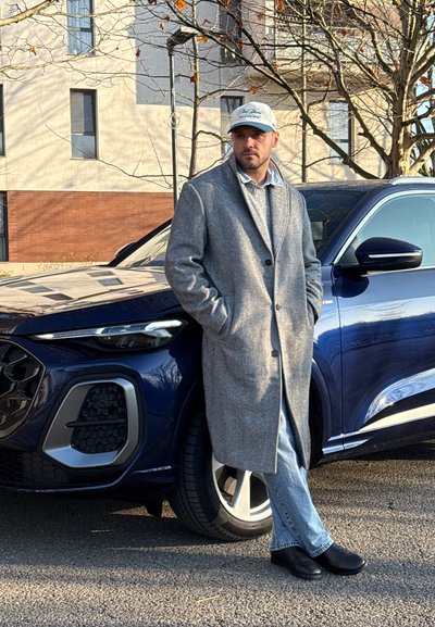Hombre con abrigo gris y gorra, con las manos en los bolsillos, apoyado contra la parte delantera de un SUV azul oscuro en una calle pavimentada.