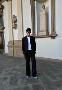 Čierny oblek s priliehavým sakom a širokými nohavicami, kombinovaný s bielou košeľou a striebornými topánkami, na neutrálnej stene.