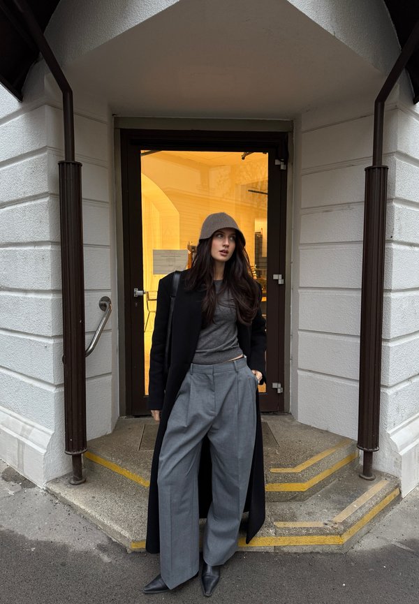 Cappotto nero sopra un top grigio, pantaloni larghi grigi e un berretto marrone. Indossati con scarpe nere a punta, in piedi accanto a una porta di vetro.