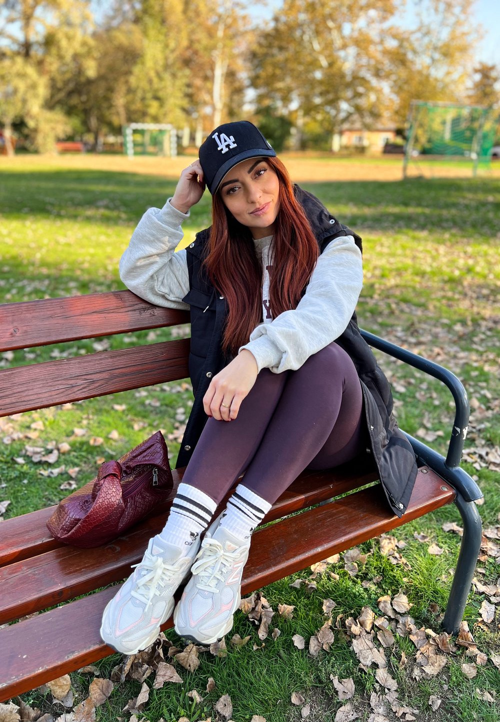 Boné preto com o logótipo da LA, sweat gris, colete preto, leggings escuras e ténis brancos com detalhes cor-de-rosa; bolsa bordô em cima do banco, folhas de outono visíveis.