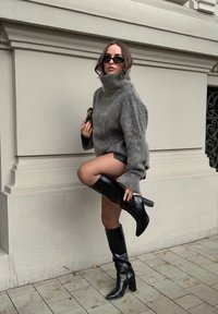 Tömör szürke pulcsi magas gallérral, fényes fekete térdizomcsizmákkal párosítva. Strukturált anyag széles ujjal, egy kis kiegészítőt tartva.