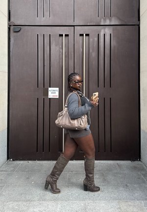 Mujer con suéter gris, pantalones cortos, botas hasta la rodilla y gafas de sol, caminando junto a una gran puerta automática marrón mientras sostiene un smartphone y un bolso beige.