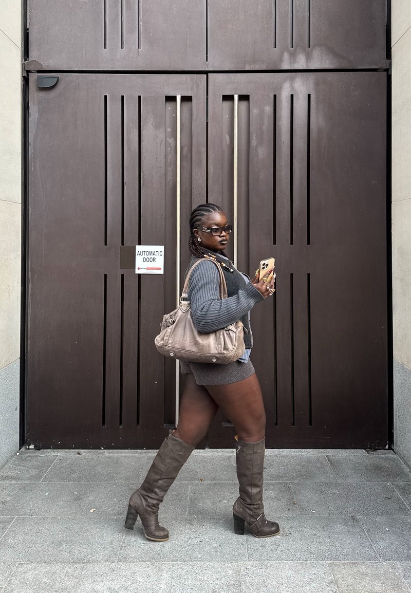 Donna che indossa un maglione grigio, pantaloncini, stivali al ginocchio e occhiali da sole, che cammina accanto a una grande porta automatica marrone, tenendo uno smartphone e una borsa beige.