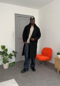 Fekete kétsoros kabát bézs pulóver felett, laza sötét farmernadrággal párosítva. Kiegészítők: fekete sapka és napszemüveg. Narancssárga szék a háttérben.