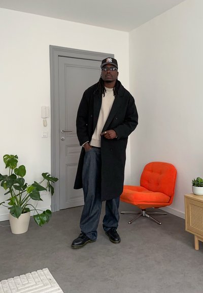 Abrigo negro cruzado sobre un suéter beige, combinado con jeans oscuros y sueltos. Los accesorios incluyen una gorra negra y gafas de sol. Silla naranja en el fondo.