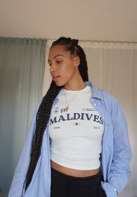 Valkoinen crop-top, jossa lukee "MALDIVEJA" tummansinisellä tekstillä, päällä vaaleansininen raidallinen nappipaita. Pitkät tummat letitetyt hiukset ja kultaketju kaulassa.