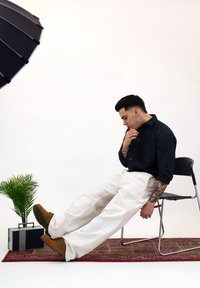 Chemise à carreaux noire, pantalon blanc à jambes larges, chaussures marron et tatouages décoratifs. Chaise et plante en pot avec une radio vintage en arrière-plan.